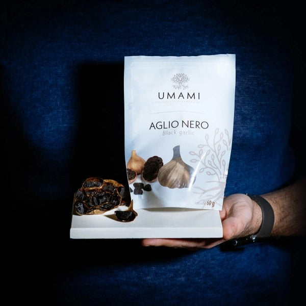 Eine Hand präsentiert einen Beutel und Knollen des schwarzen Knoblauchs von Umami